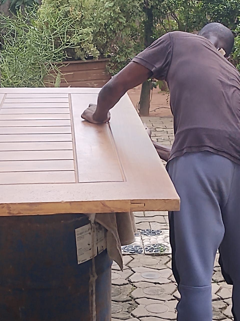 Artisan ponçant une porte en bois clair posée sur un baril bleu en extérieur.