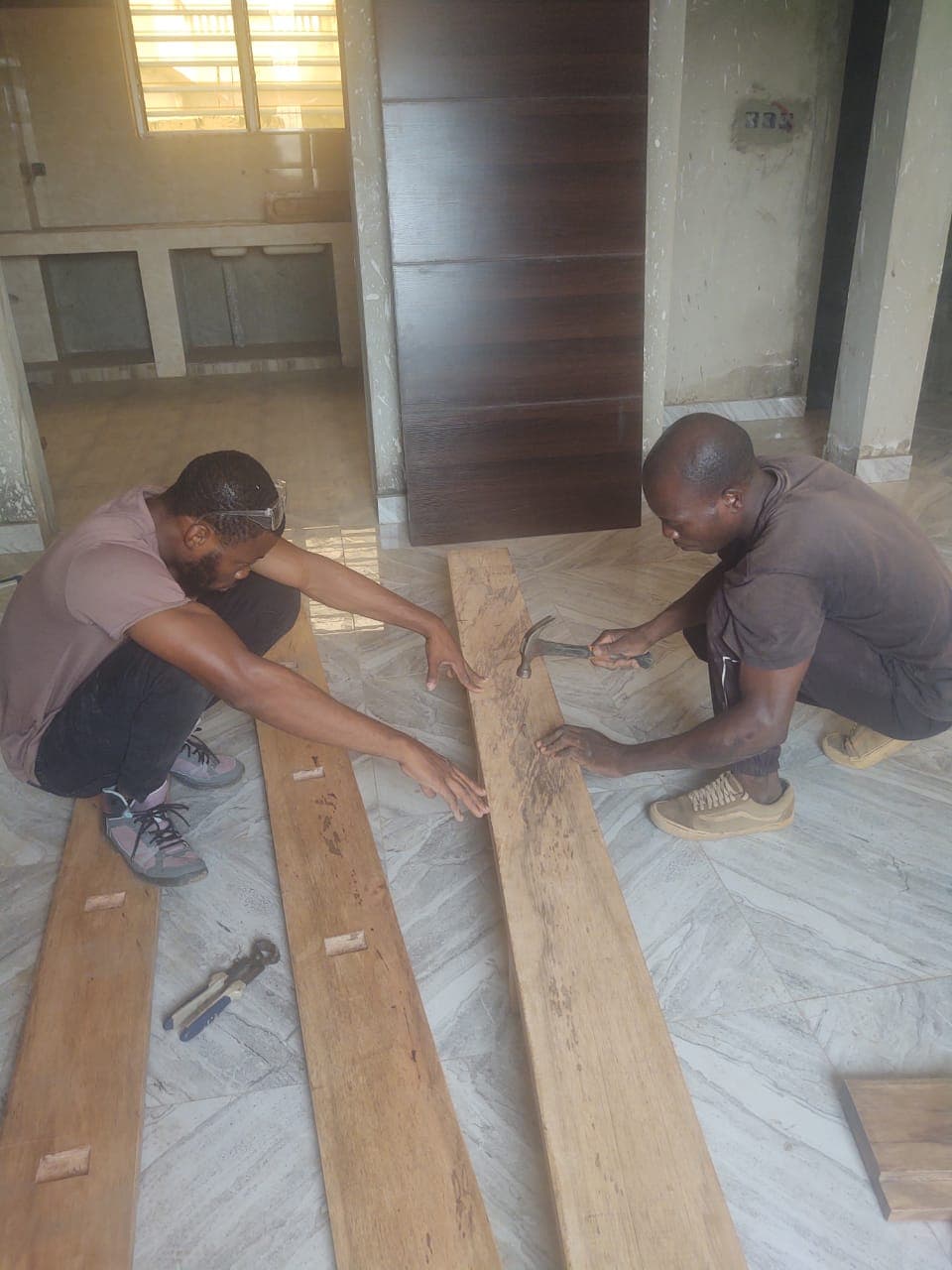 Deux ouvriers accroupis assemblent des planches de bois avec un marteau sur un sol carrelé.