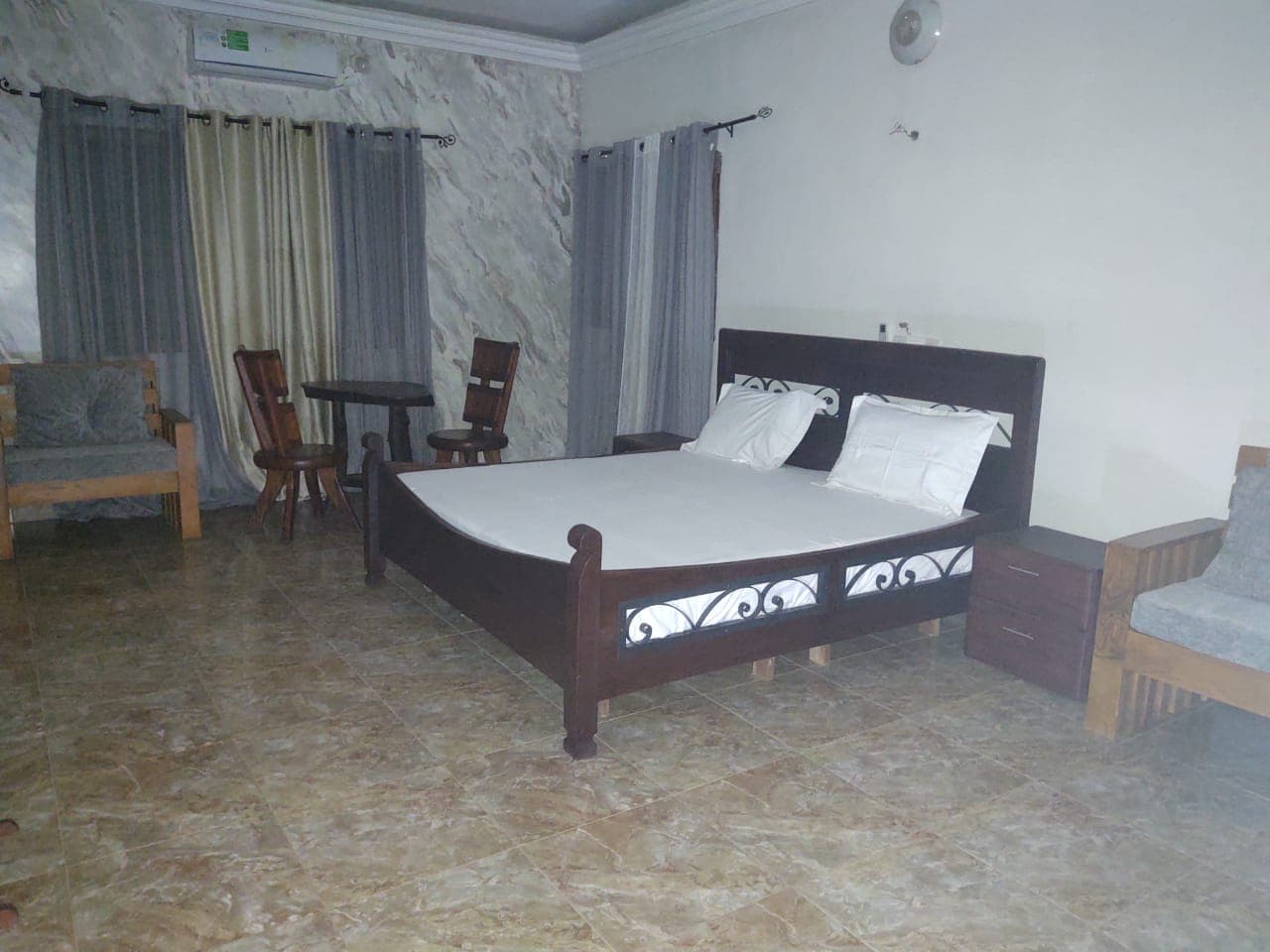 Chambre avec lit en bois sombre, draps blancs, carrelage beige, rideaux gris et mobilier assorti.